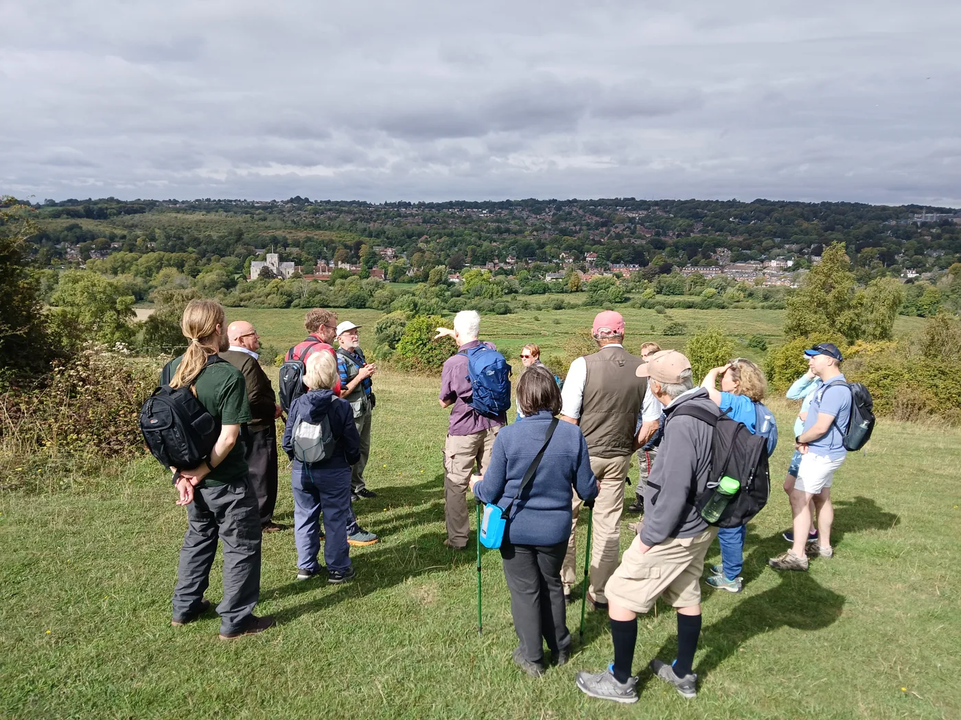 A group of walkers standing on a grassy hillside, with trees and houses across the valley behind them